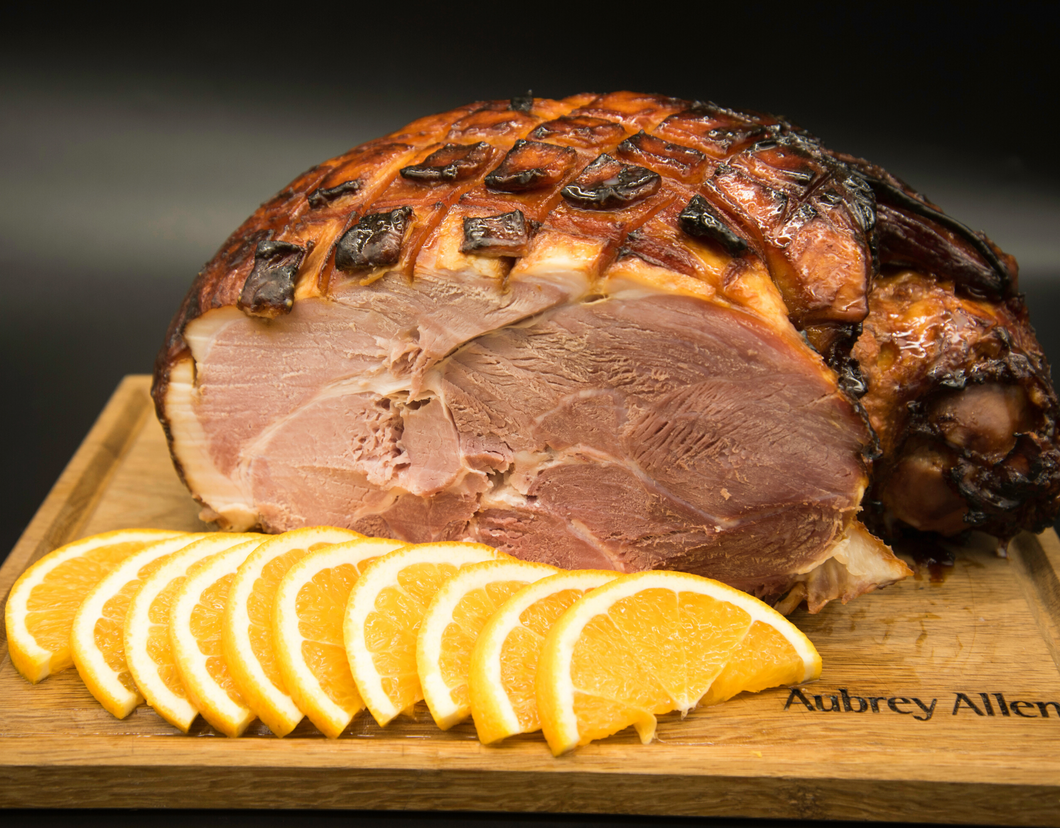 A whole cooked ham, honey glazed top, on a board with slices of orange